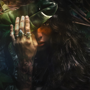 Green witch portrait of woman with black hair, jewellery tattoos behind leaves
